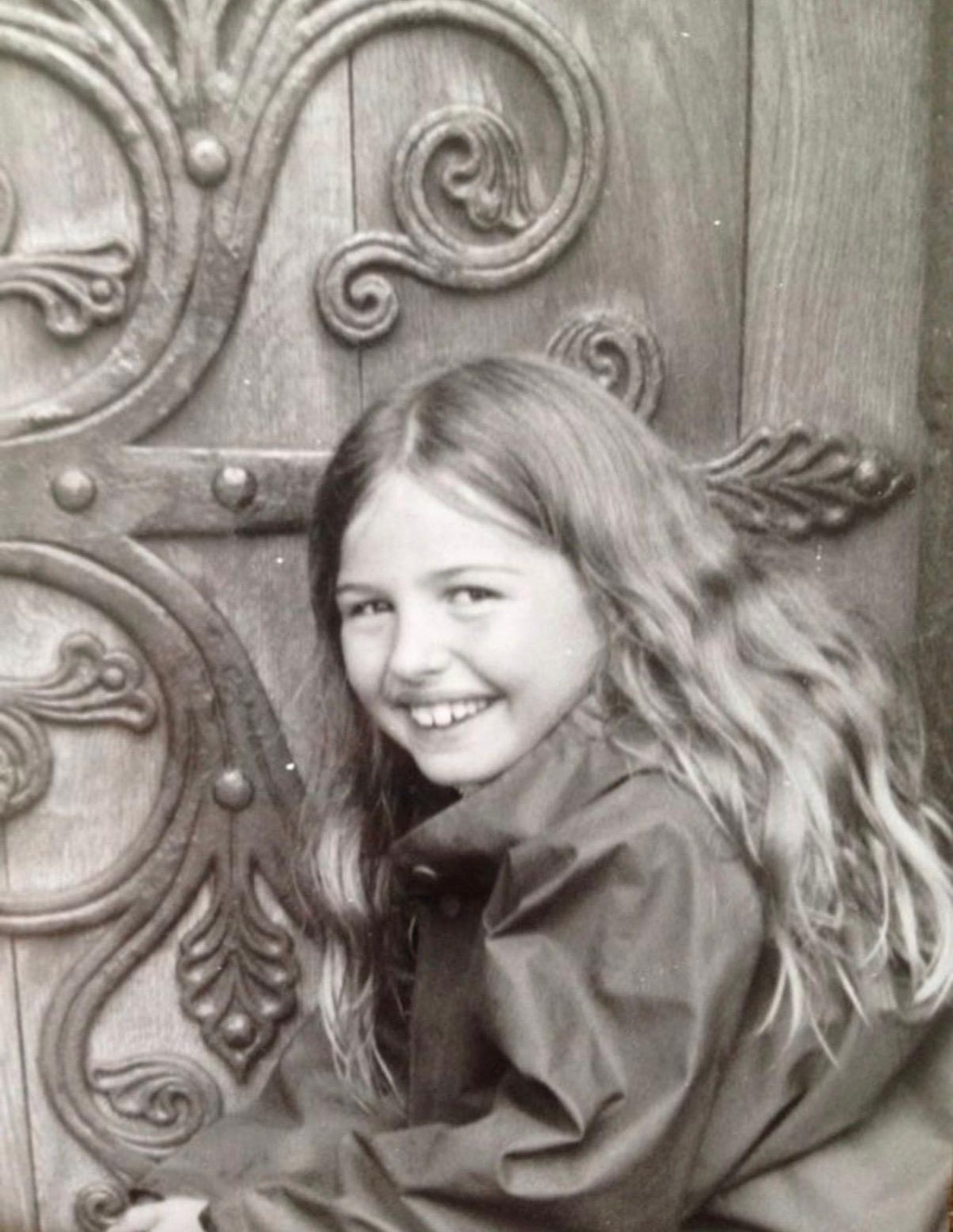 Black-and-white childhood photo: Marieke McKenna as a girl, smiling over her shoulder in front of a carved wooden door with iron scrollwork