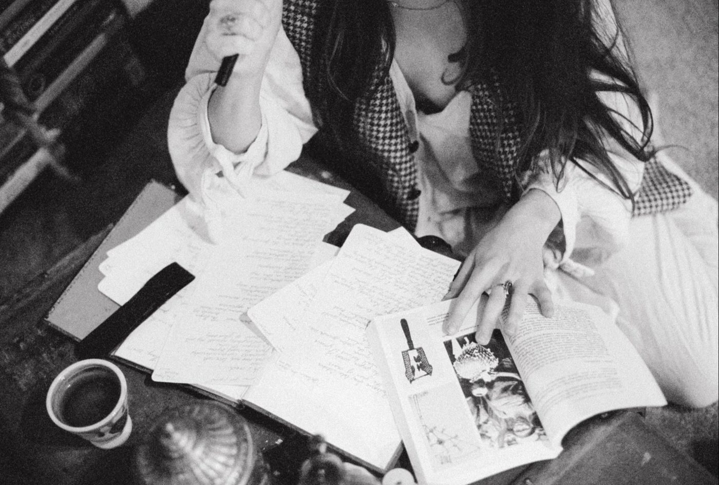 Seen from above: Marieke McKenna at a table covered in handwritten pages and an open book, pen in hand, a cup of coffee beside her