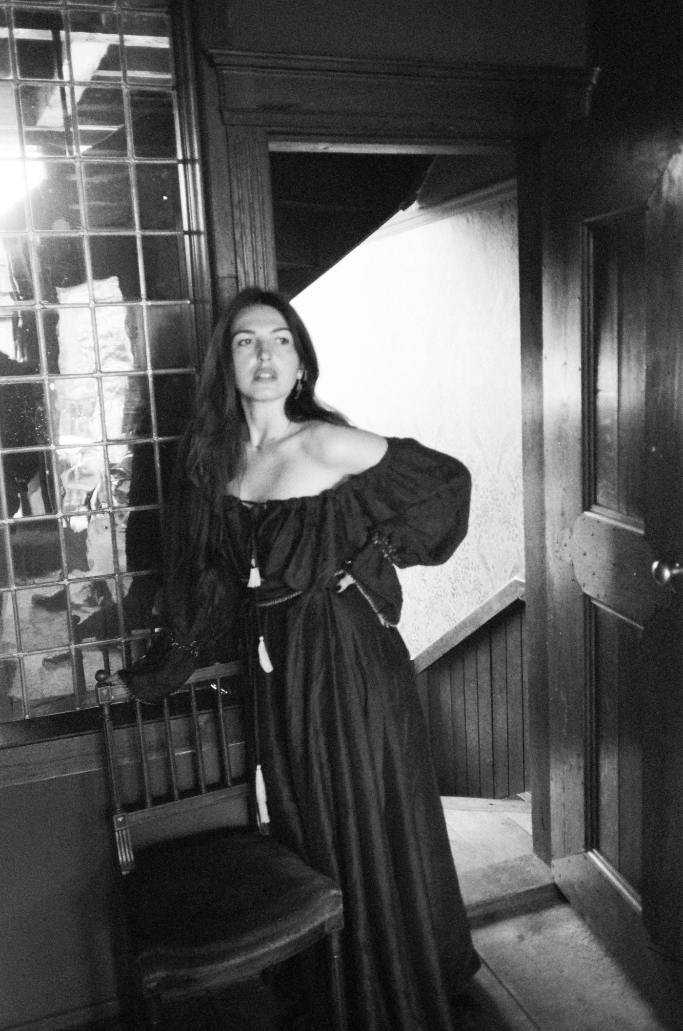 Marieke McKenna in a long black off-shoulder dress by a leaded window and an open wooden door