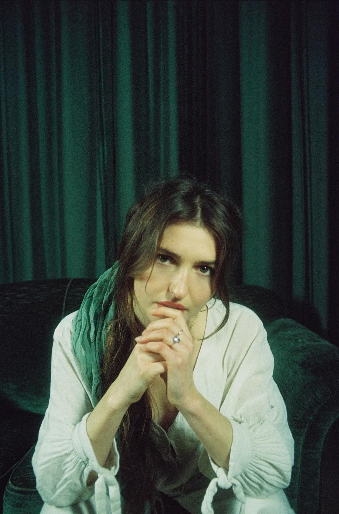 Marieke McKenna seated in a green velvet armchair before a green curtain, hands clasped, looking into the camera