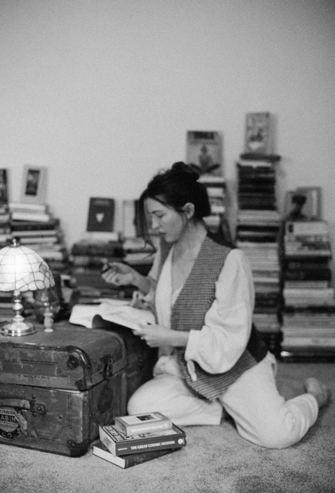 Marieke McKenna reading on the floor among stacks of books, beside an old travel trunk and a lamp