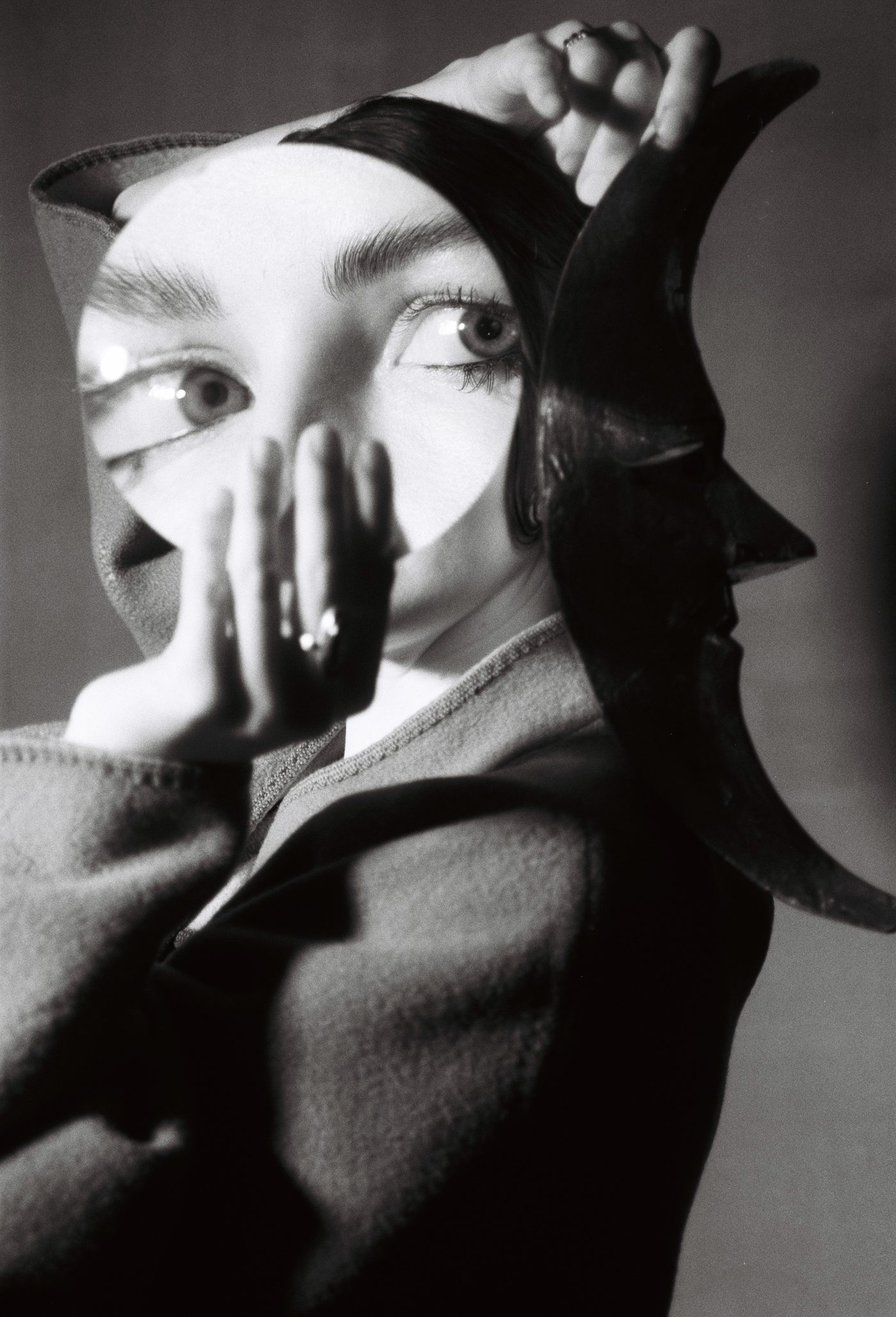 Marieke McKenna holding a round mirror and a carved crescent-moon mask to her face, one eye doubled in the mirror
