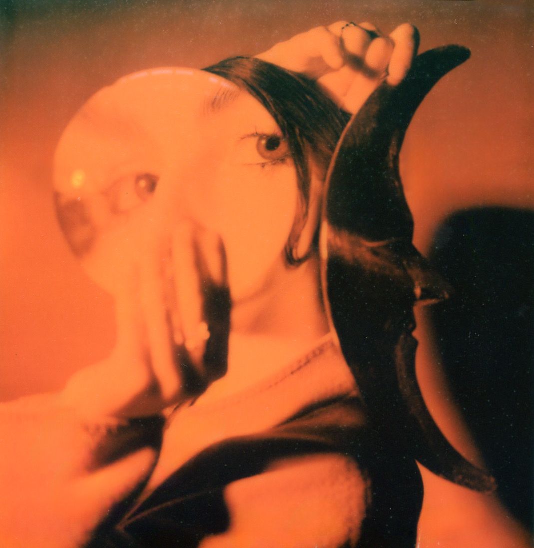 An orange Polaroid of Marieke McKenna with a mirror and a crescent-moon mask