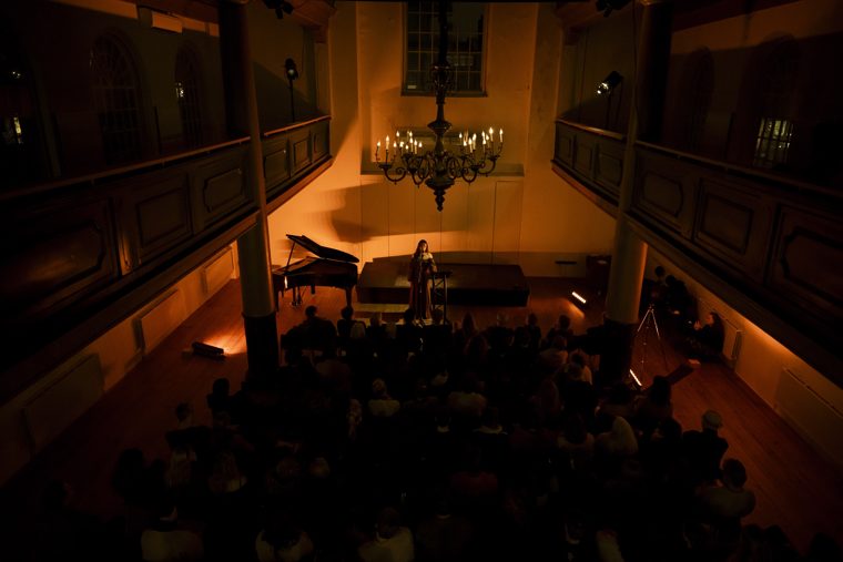 The Uilenburgersjoel from the balcony: Marieke McKenna at a microphone beside a grand piano under a candle chandelier, before a full audience