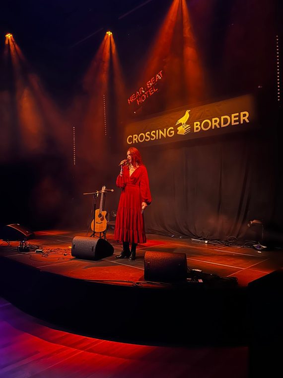 Marieke McKenna in a red dress presenting on the Crossing Border stage in red and amber light