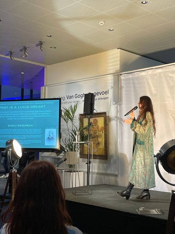 Marieke McKenna on stage with a microphone before Van Gogh’s Sunflowers and a screen asking What is a lucid dream?